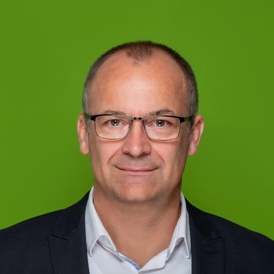 Portr&auml;t eines Mannes in Anzug mit Brille vor gr&uuml;nem Hintergrund, freundlich l&auml;chelnd.