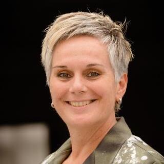 Portrait einer l&auml;chelnden Frau mit kurzem, blondem Haar, die einen grauen Blazer mit Mustern tr&auml;gt.