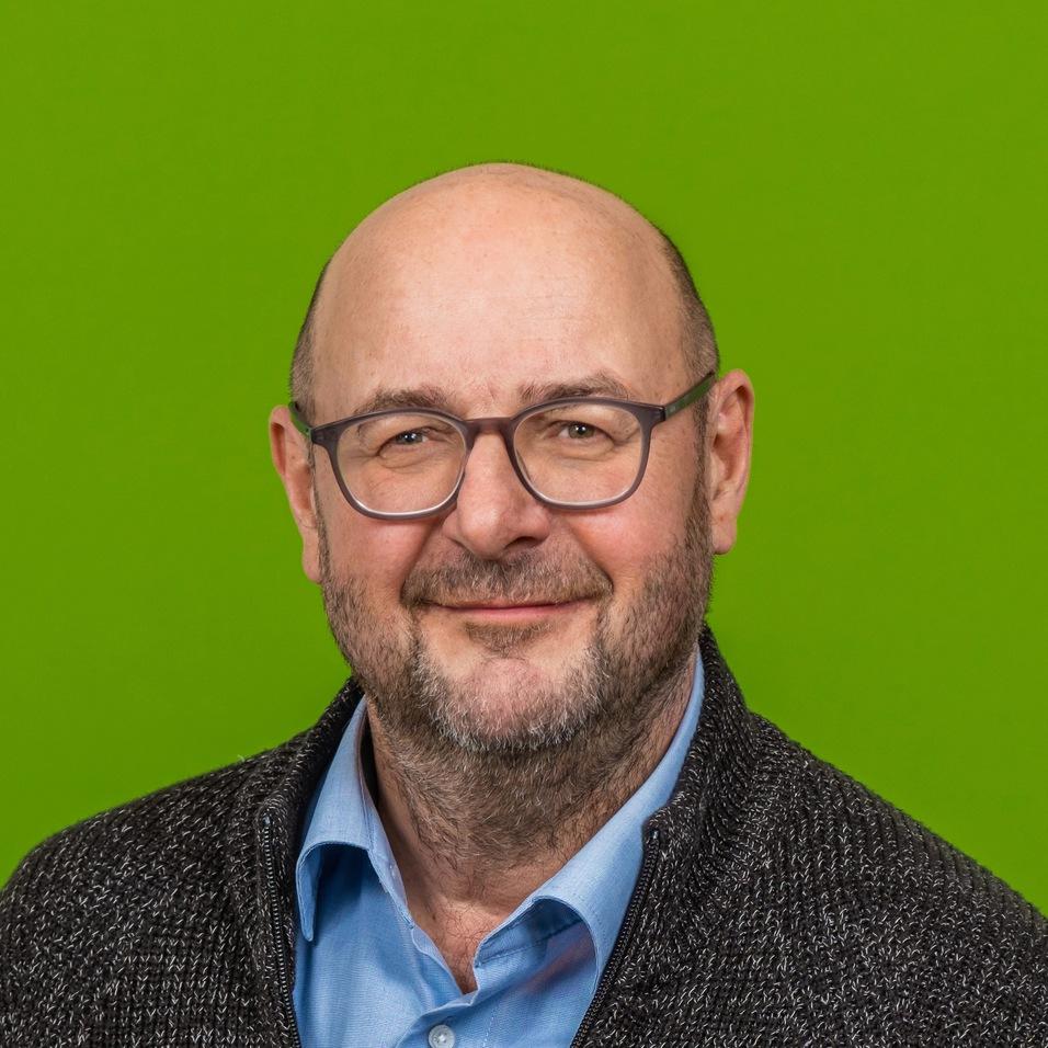 Mann mit Brille und kurzem Bart l&auml;chelt vor gr&uuml;nem Hintergrund, tr&auml;gt einen schwarzen Pullover und ein hellblaues Hemd.