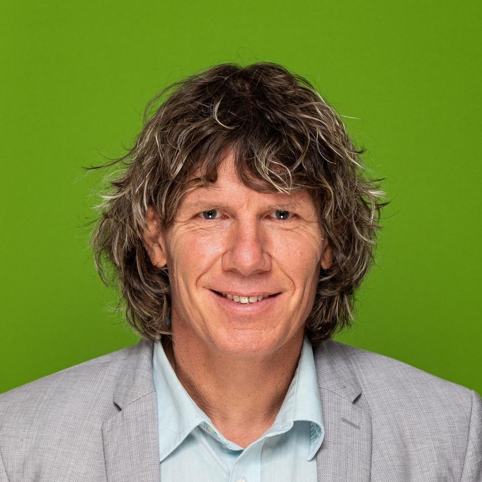 Porträt eines Mannes mit lockigem Haar, der einen grauen Anzug und ein hellblaues Hemd trägt, vor grünem Hintergrund.