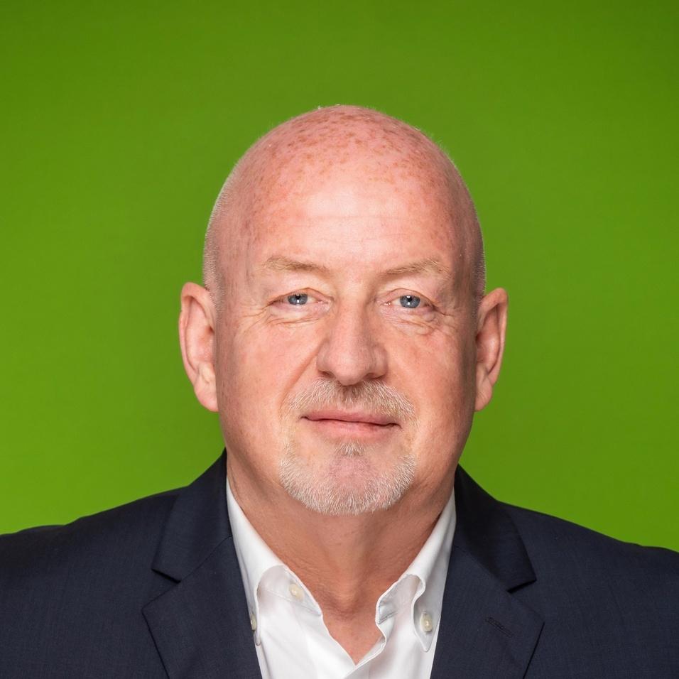 Porträt eines Mannes mit Glatze in Anzug vor grünem Hintergrund, blickt neutral in die Kamera.