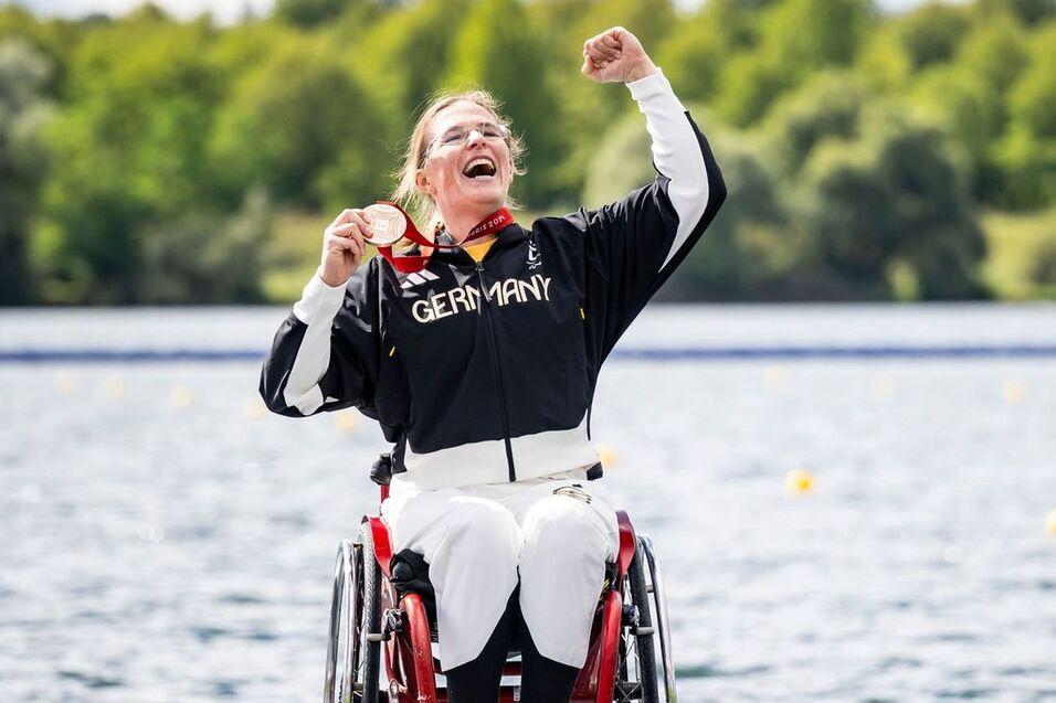 Eine Frau im Rollstuhl jubelt auf dem Siegerpodest und h&auml;lt eine Medaille, w&auml;hrend ein Mann im Hintergrund steht.