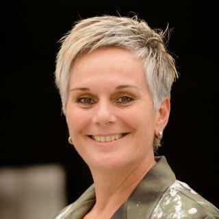 Portrait einer lächelnden Frau mit kurzem, blondem Haar, die einen grauen Blazer mit Mustern trägt.