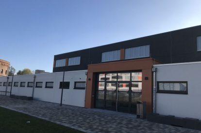 Moderne Geb&auml;udefront mit gro&szlig;en Fenstern und Eingangsbereich, umgeben von einer gepflasterten Fl&auml;che und gr&uuml;nem Gras.