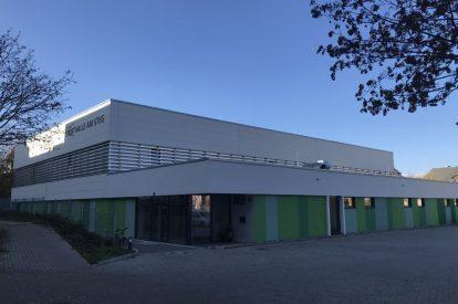 Moderne Sporthalle mit grauer Fassade und gr&uuml;nen Elementen, umgeben von einem gepflasterten Platz und B&auml;umen.