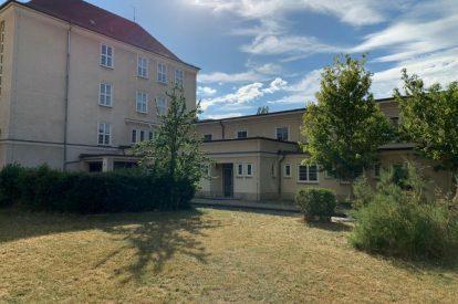 Ansicht eines Gebäudes mit zwei Etagen, umgeben von Rasenfläche und Bäumen, unter einem blauen Himmel mit Wolken.