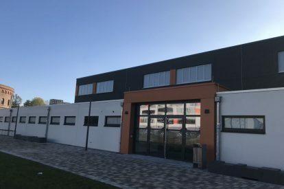 Moderne Gebäudefront mit großen Fenstern und Eingangsbereich, umgeben von einer gepflasterten Fläche und grünem Gras.