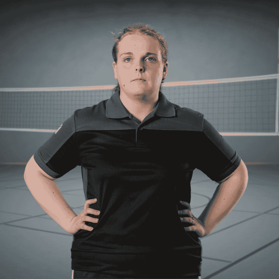 Porträt einer sportlichen Person in schwarzem Polo, stehend vor einem Volleyballnetz in einer Halle.