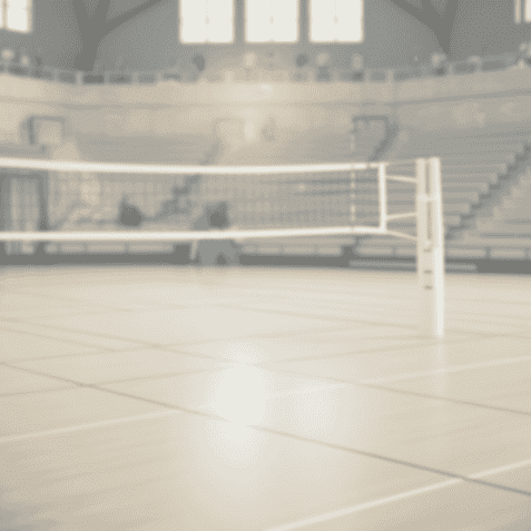 Volleyballfeld mit Netz in einer Sporthalle, leere Tribünen und diffuse Beleuchtung im Hintergrund.