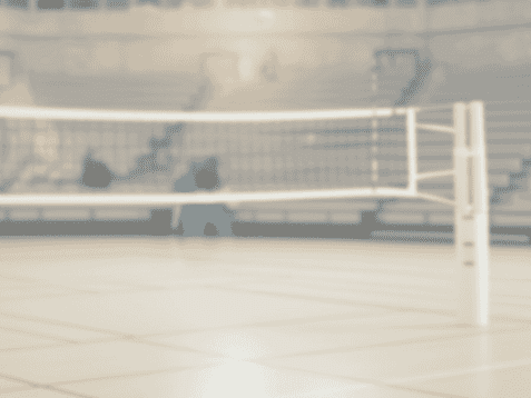 Volleyballfeld mit Netz in einer Sporthalle, leere Tribünen und diffuse Beleuchtung im Hintergrund.