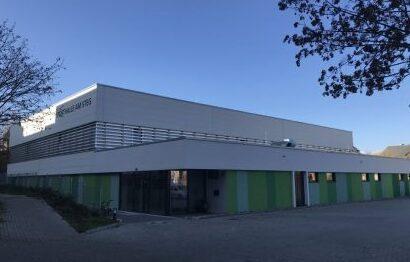 Moderne Sporthalle mit grauer Fassade und gr&uuml;nen Elementen, umgeben von einem gepflasterten Platz und B&auml;umen.