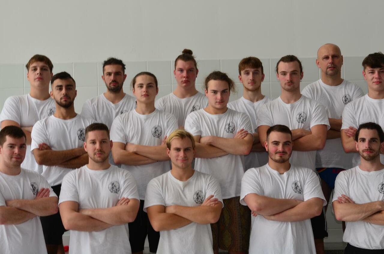 Gruppenfoto von 15 männlichen Sportlern in weißen T-Shirts, stehend und mit verschränkten Armen auf einem Sportgelände.