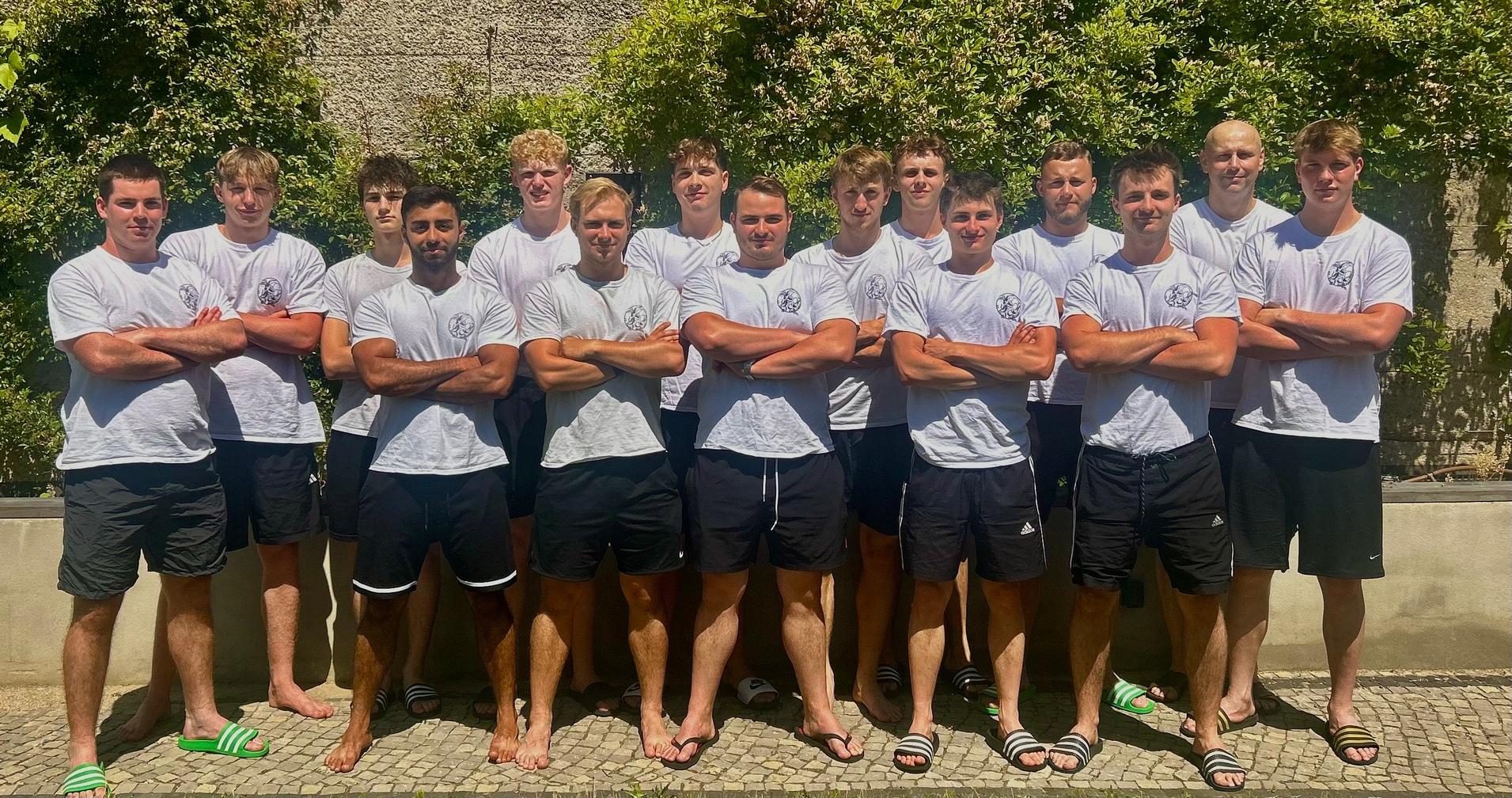 Gruppe von zwölf jungen Männern in weißen T-Shirts und schwarzen Shorts, posed vor einer grünen Hecke im Freien.