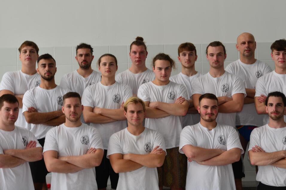 Gruppenfoto von 15 männlichen Sportlern in weißen T-Shirts, stehend und mit verschränkten Armen auf einem Sportgelände.
