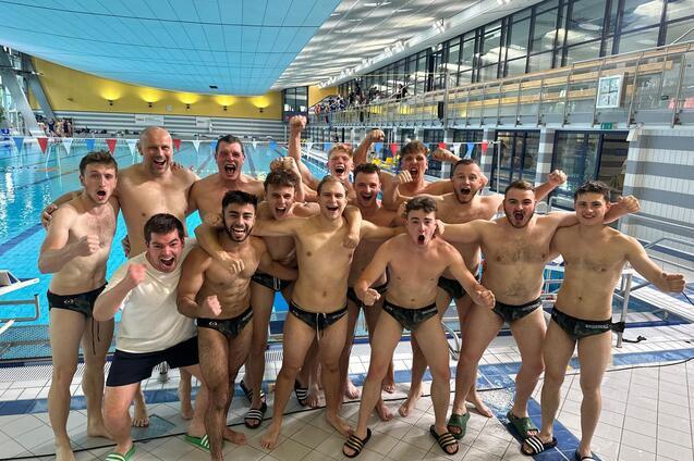 Gruppe junger M&auml;nner in Badeanz&uuml;gen posiert jubelnd am Beckenrand eines Schwimmbades.