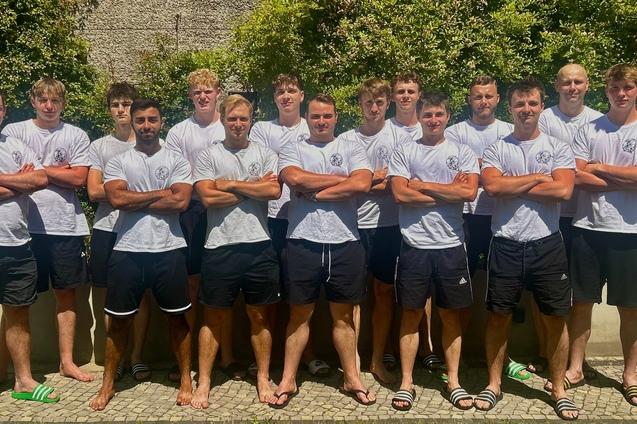 Gruppe von zw&ouml;lf jungen M&auml;nnern in wei&szlig;en T-Shirts und schwarzen Shorts, posed vor einer gr&uuml;nen Hecke im Freien.