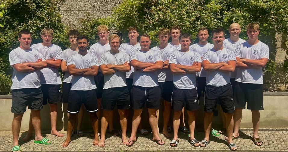 Gruppe von zwölf jungen Männern in weißen T-Shirts und schwarzen Shorts, posed vor einer grünen Hecke im Freien.