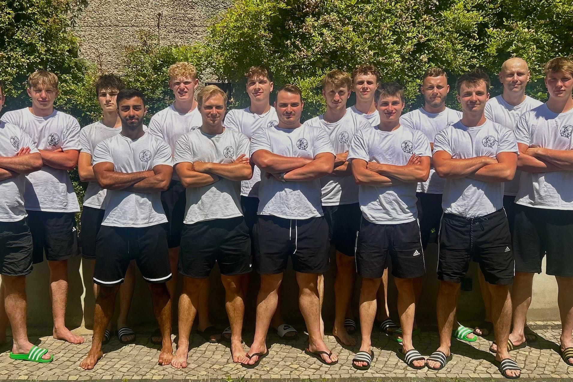 Gruppe von zw&ouml;lf jungen M&auml;nnern in wei&szlig;en T-Shirts und schwarzen Shorts, posed vor einer gr&uuml;nen Hecke im Freien.