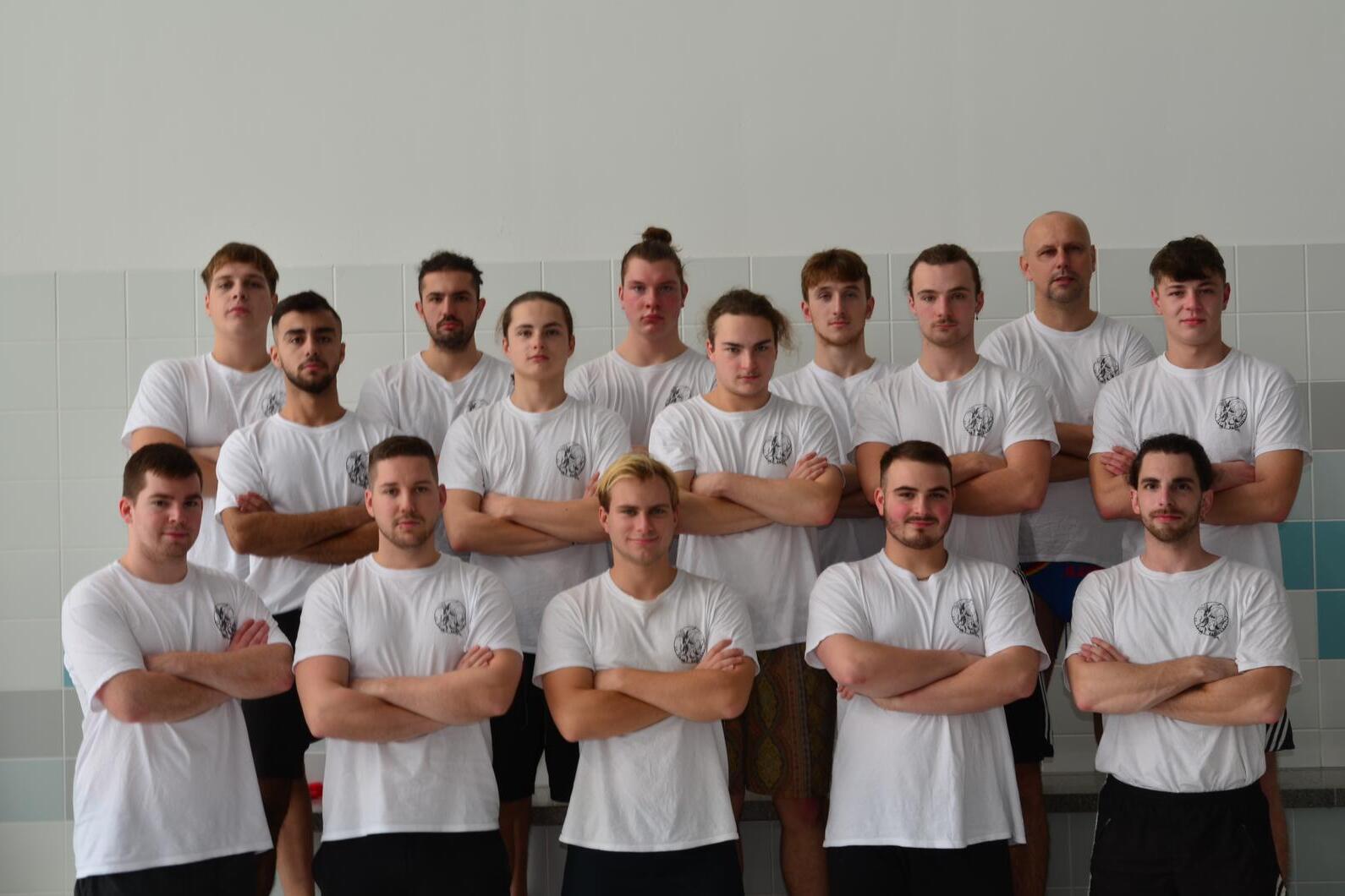 Gruppenfoto von 15 m&auml;nnlichen Sportlern in wei&szlig;en T-Shirts, stehend und mit verschr&auml;nkten Armen auf einem Sportgel&auml;nde.