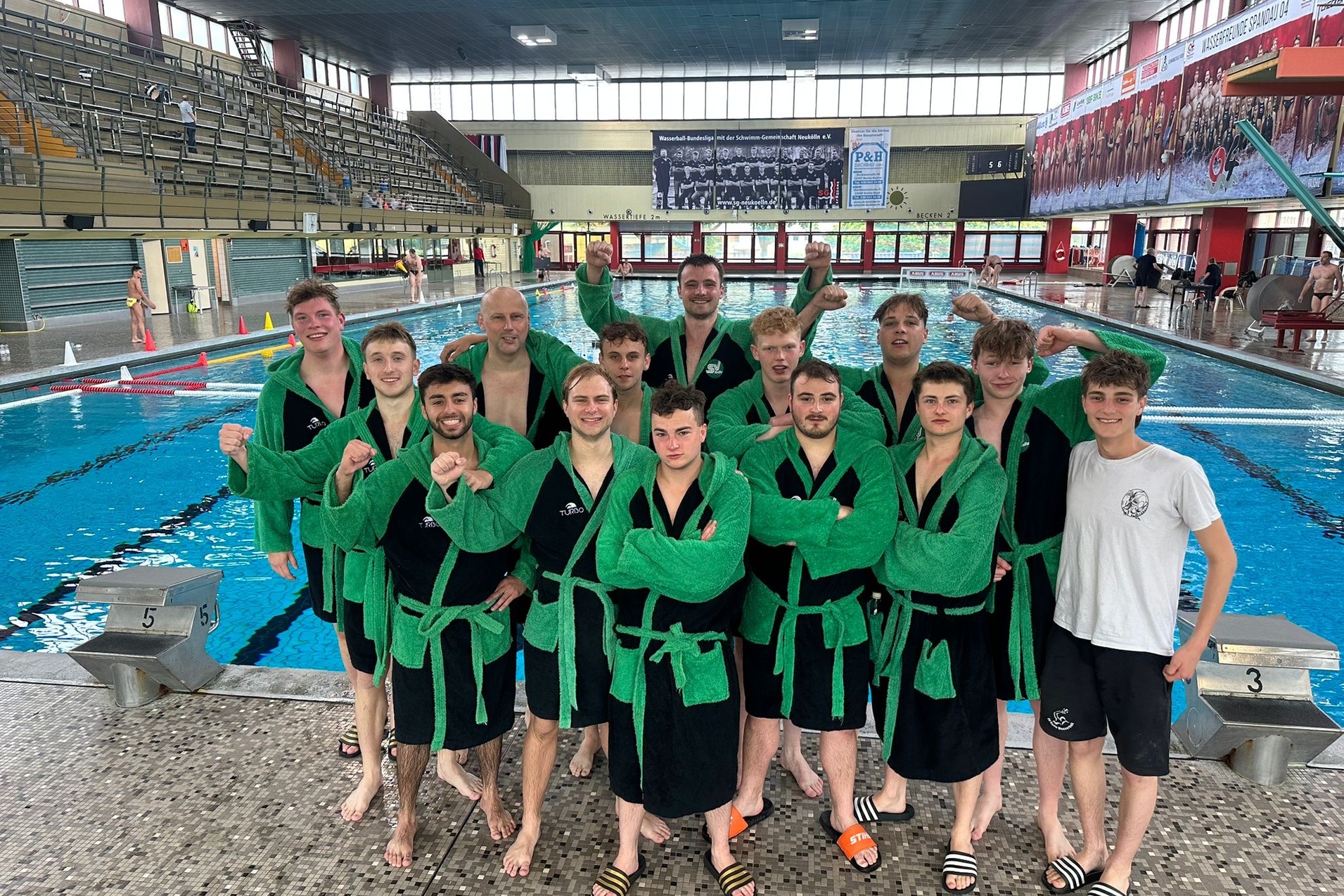 Gruppe von Schwimmern in gr&uuml;nen Badekappen und -m&auml;nteln, posierend am Beckenrand eines Schwimmbads.