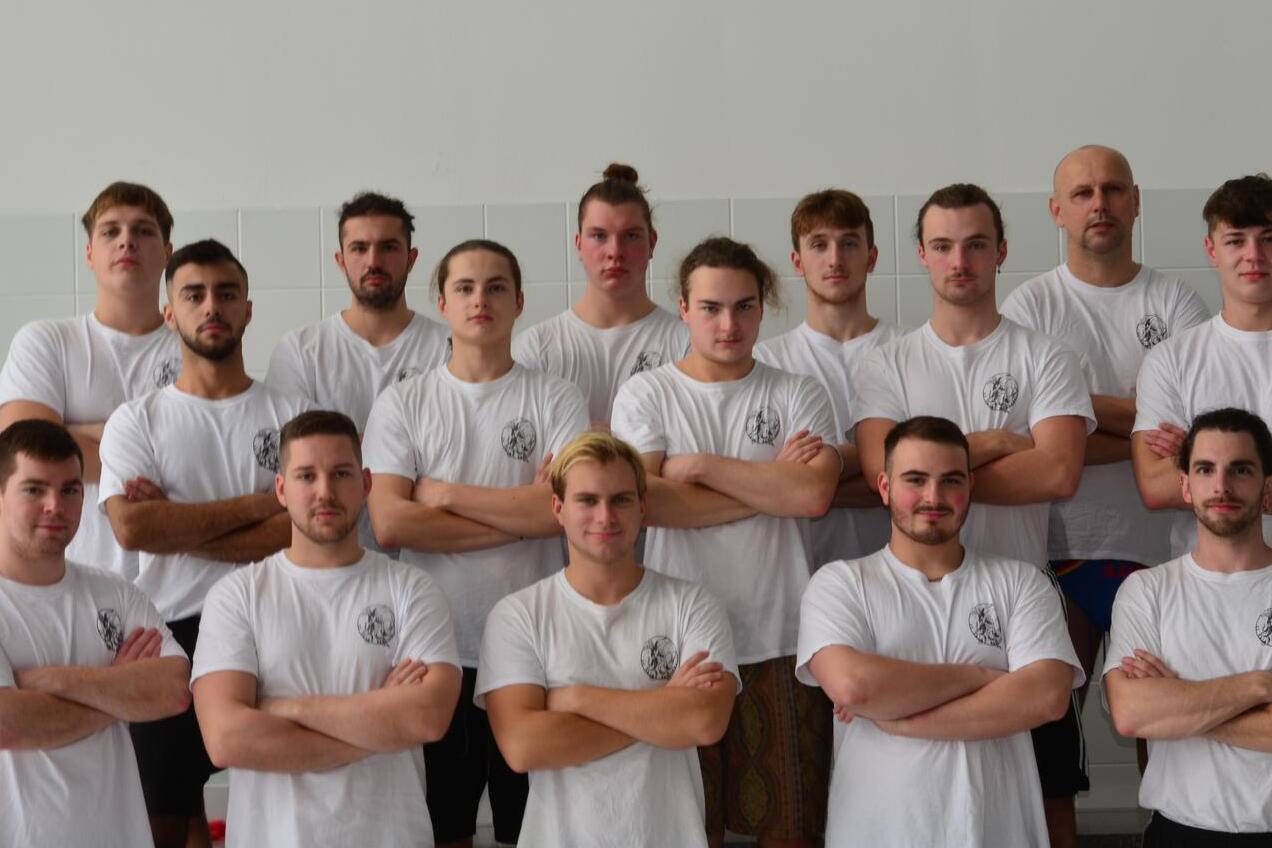 Gruppenfoto von 15 männlichen Sportlern in weißen T-Shirts, stehend und mit verschränkten Armen auf einem Sportgelände.