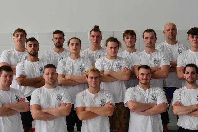 Gruppenfoto von 15 m&auml;nnlichen Sportlern in wei&szlig;en T-Shirts, stehend und mit verschr&auml;nkten Armen auf einem Sportgel&auml;nde.
