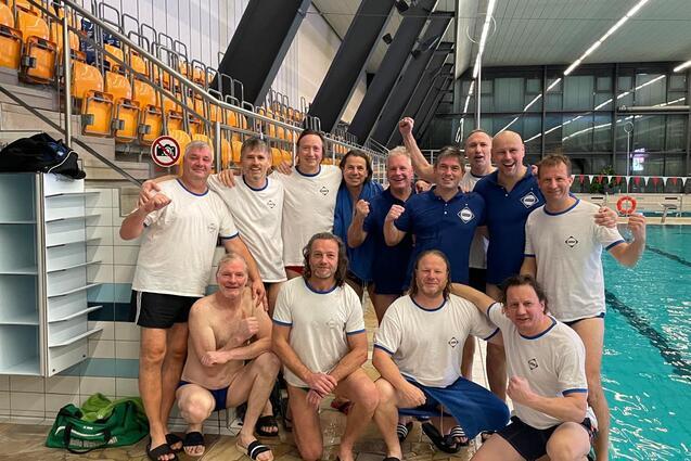 Gruppe von Schwimmern in Badebekleidung posiert vor einem Pool, l&auml;chelnd und die Arme hochgehend.