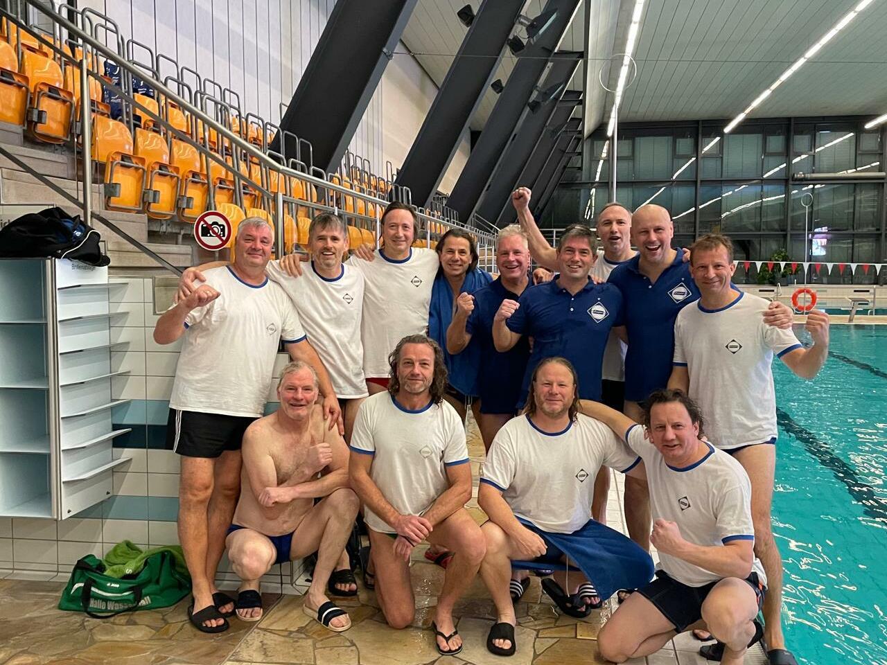 Gruppe von Schwimmern in Badebekleidung posiert vor einem Pool, lächelnd und die Arme hochgehend.