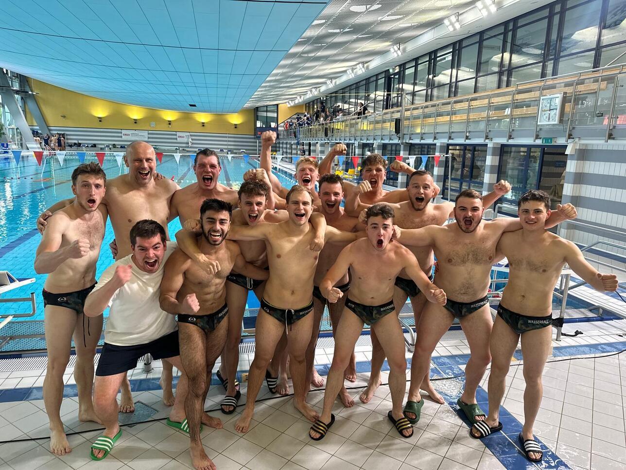 Gruppe junger Männer in Badeanzügen posiert jubelnd am Beckenrand eines Schwimmbades.