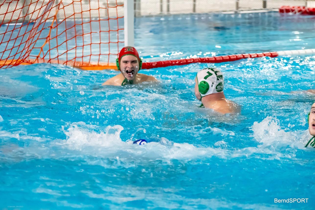 Zwei Wasserballspieler in einem Becken, einer lacht, während der andere mit einem Ball schwimmt. Rote Tor-Netz im Hintergrund.