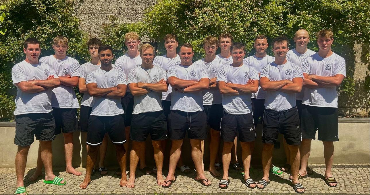 Gruppe von zwölf jungen Männern in weißen T-Shirts und schwarzen Shorts, posed vor einer grünen Hecke im Freien.