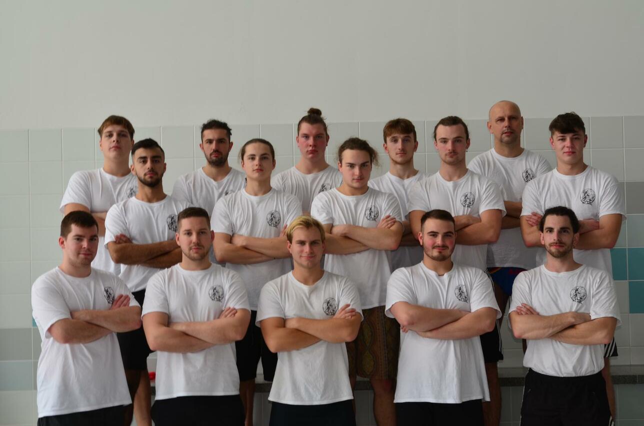 Gruppenfoto von 15 männlichen Sportlern in weißen T-Shirts, stehend und mit verschränkten Armen auf einem Sportgelände.