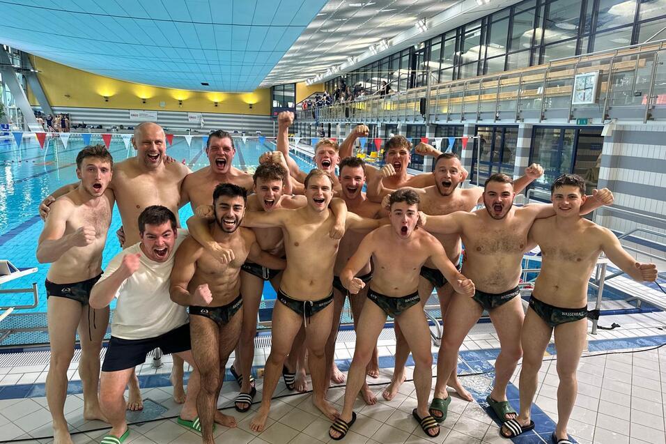 Gruppe junger Männer in Badeanzügen posiert jubelnd am Beckenrand eines Schwimmbades.