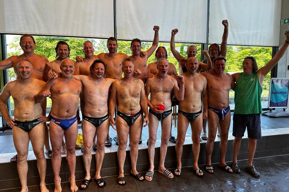 Gruppe von 15 Männern in Badebekleidung posiert fröhlich vor Fenster mit hochgestreckten Armen und Wasserball.