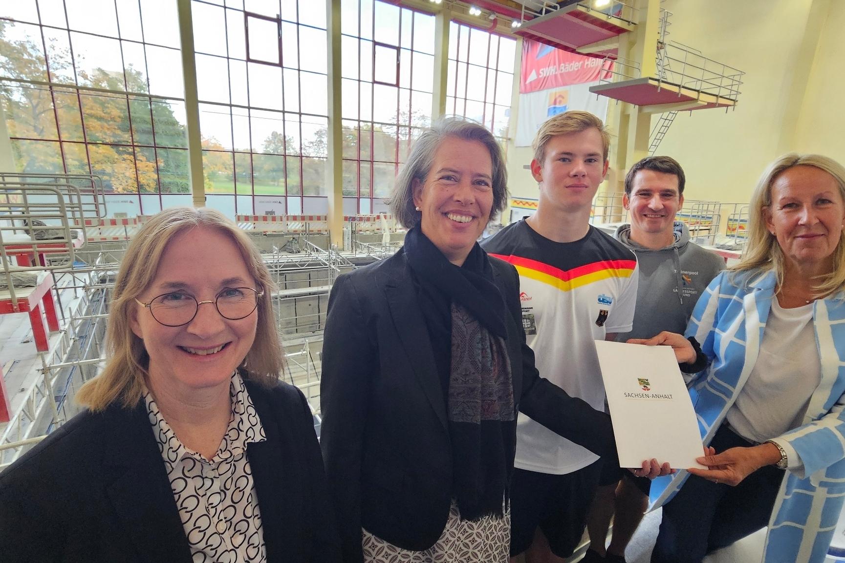 Fördermittel Sprunghalle Gruppenfoto mit fünf Personen in einem Schwimmbad, eine Person hält ein Dokument mit dem Logo „Sachsen-Anhalt“.