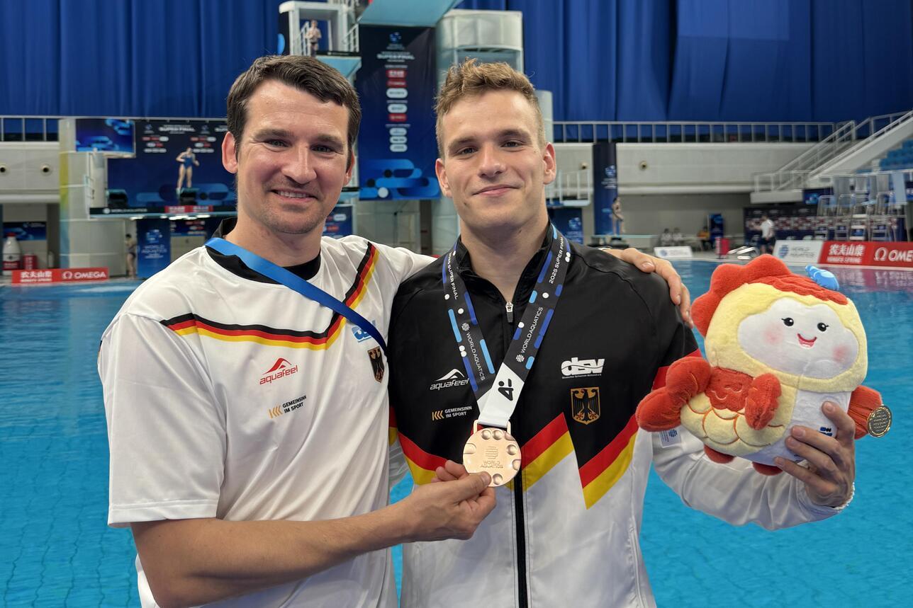 Zwei Männer in Sportkleidung stehen am Beckenrand und halten eine Medaille sowie ein Stofftier in der Hand.