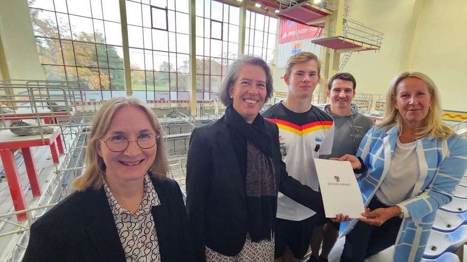 Fördermittel Sprunghalle Gruppenfoto mit fünf Personen in einem Schwimmbad, eine Person hält ein Dokument mit dem Logo „Sachsen-Anhalt“.