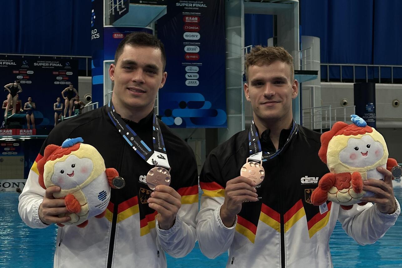 Zwei Sportler in deutscher Kleidung halten Medaillen und Plüschtiere vor einem Wettbewerbspool.