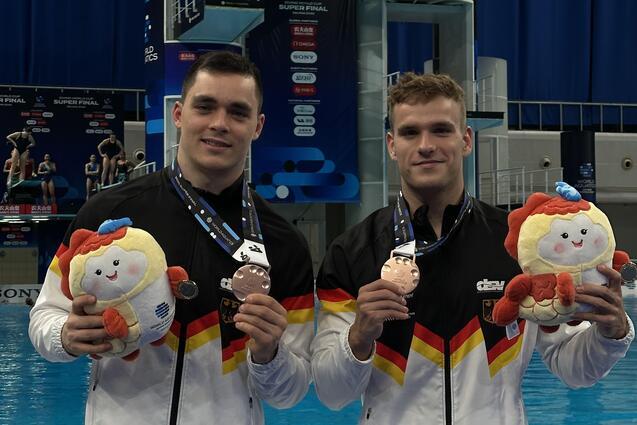 Zwei Sportler in deutscher Kleidung halten Medaillen und Pl&uuml;schtiere vor einem Wettbewerbspool.