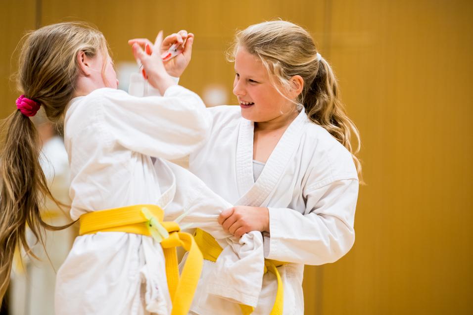 Zwei Mädchen in weißen Karateanzügen mit gelben Gurten üben freundschaftlich im Trainingsraum miteinander.