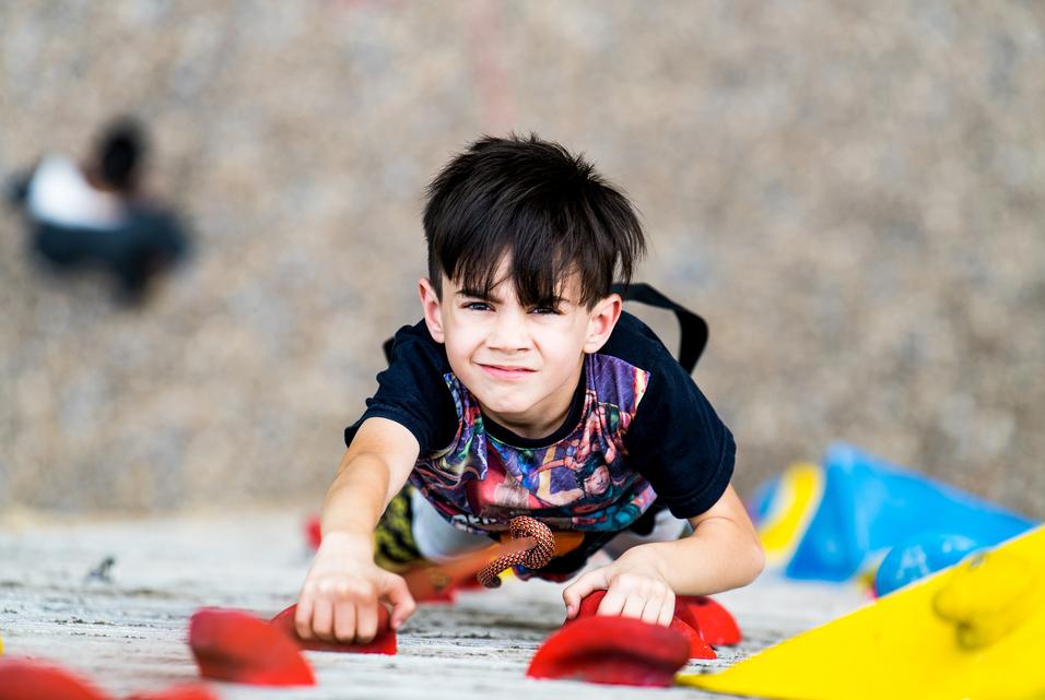 Junge klettert an einer Bouldering-Wand, greift nach einem roten Griff, mit angespannter Miene und sportlicher Kleidung.