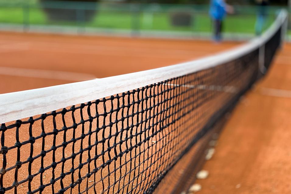 Nahaufnahme eines Tennisnetzes auf einem roten Sandplatz, Hintergrund unscharf mit grünen Bäumen.