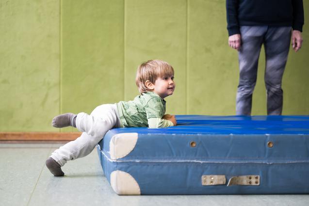 Kleines Kind krabbelt auf einer blauen Turnmatte in einer Turnhalle mit grünen Wänden.