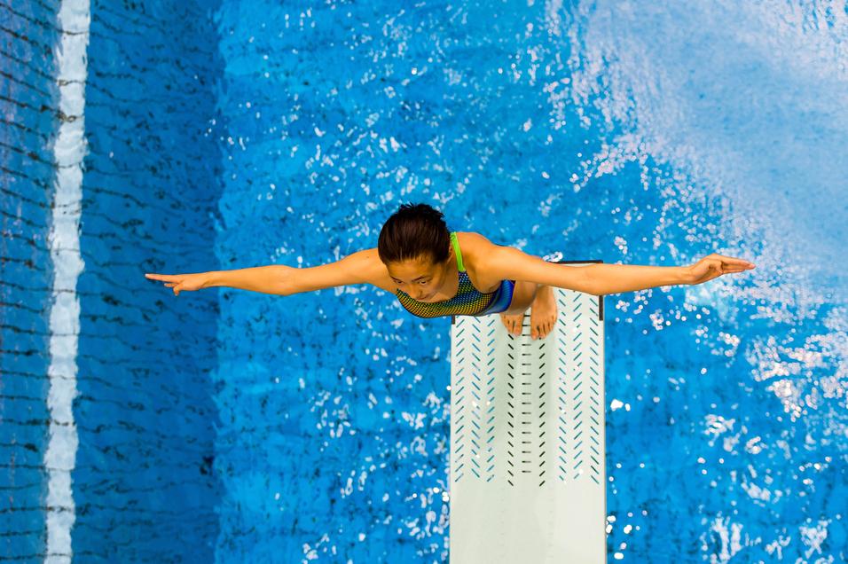 Junge Sportlerin steht auf einem Sprungbrett, bereit zum Sprung in einem blauen Schwimmbecken.